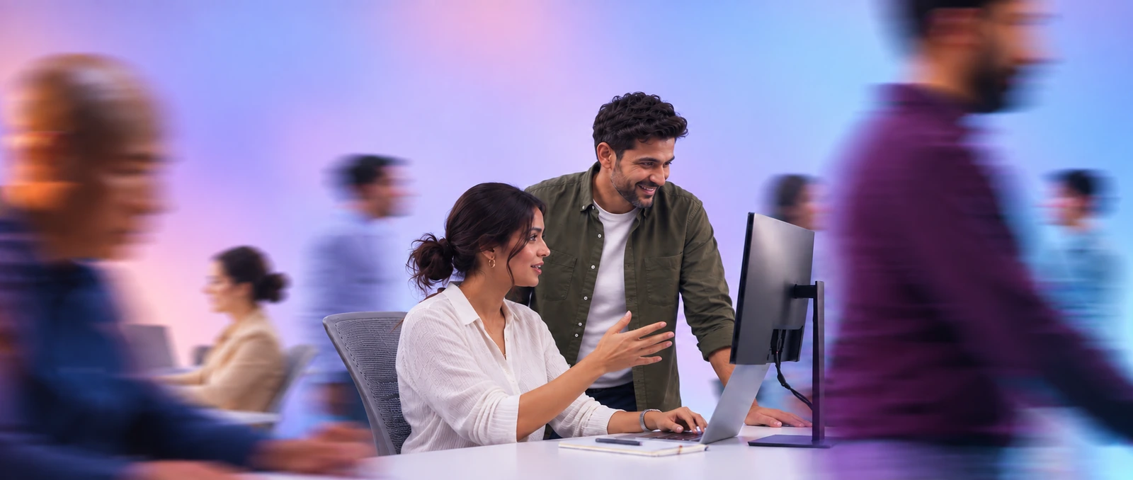 Two colleagues at a desk reading the same screen, the office moving past behind them